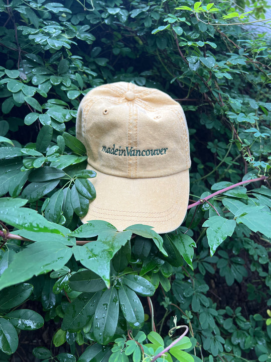 Mustard pigment-dyed cap with 'made in Vancouver' embroidered text on a green leafy background