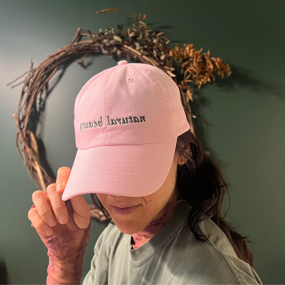 Person wearing the Cascadia Natural Beauty cap, standing in front of a decorative wreath on a green wall.
